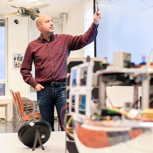 Vorlesung Auf dem Bild ist ein Professor zu sehen. Er zeigt auf eine interaktive Tafel. Vorne ist unscharf ein Satellitenmodell zu erkennen.