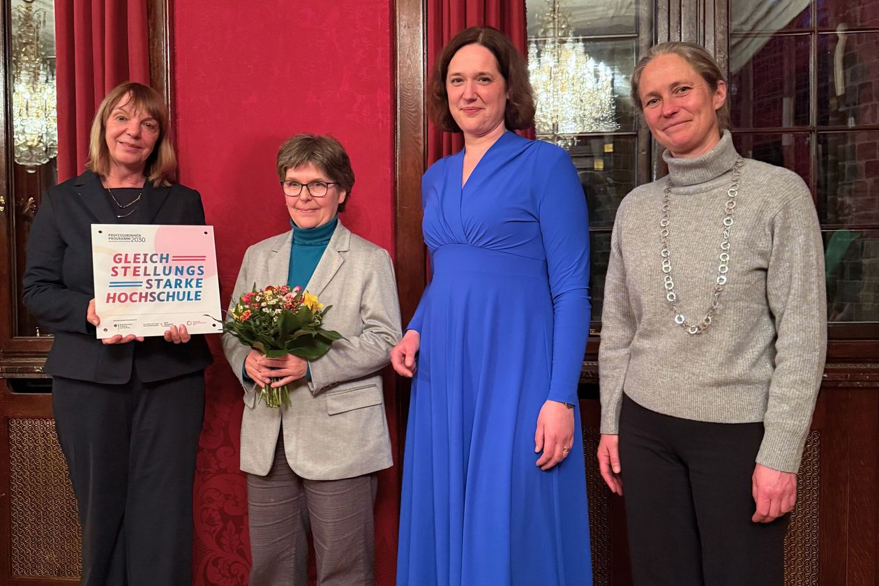 Auf dem Bild sind vier Frauen in einer Reihe aufgestellt. Die linken beiden Frauen halten ein Schild und einen Blumenstrauß in den Händen. Auf dem Schild steht: Gleichstellungsstarke Hochschule. 