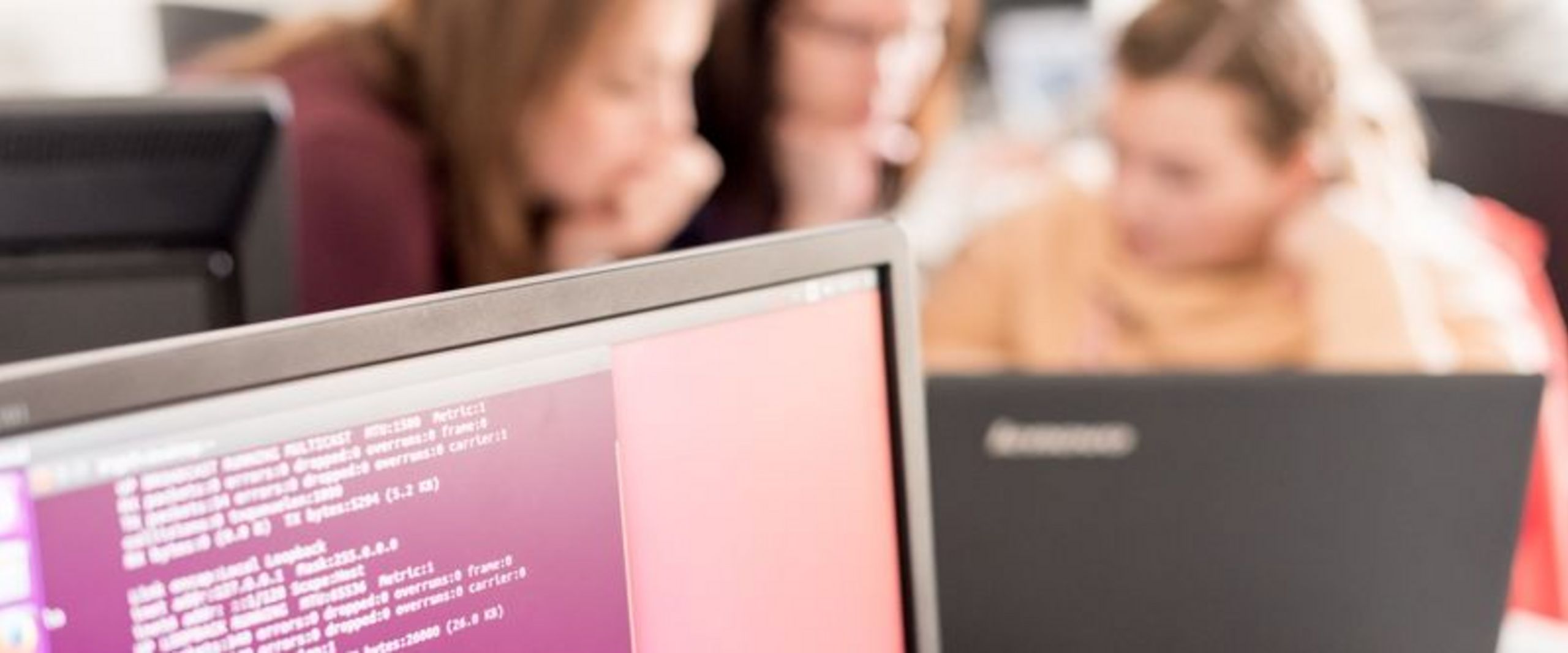 Bildschirm mit Codezeilen im Vordergrund, dahinter unscharf drei Studentinnen, die an einem Laptop sitzen und programmieren.