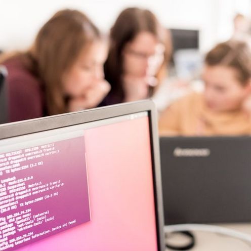 Bildschirm mit Codezeilen im Vordergrund, dahinter unscharf drei Studentinnen, die an einem Laptop sitzen und programmieren.