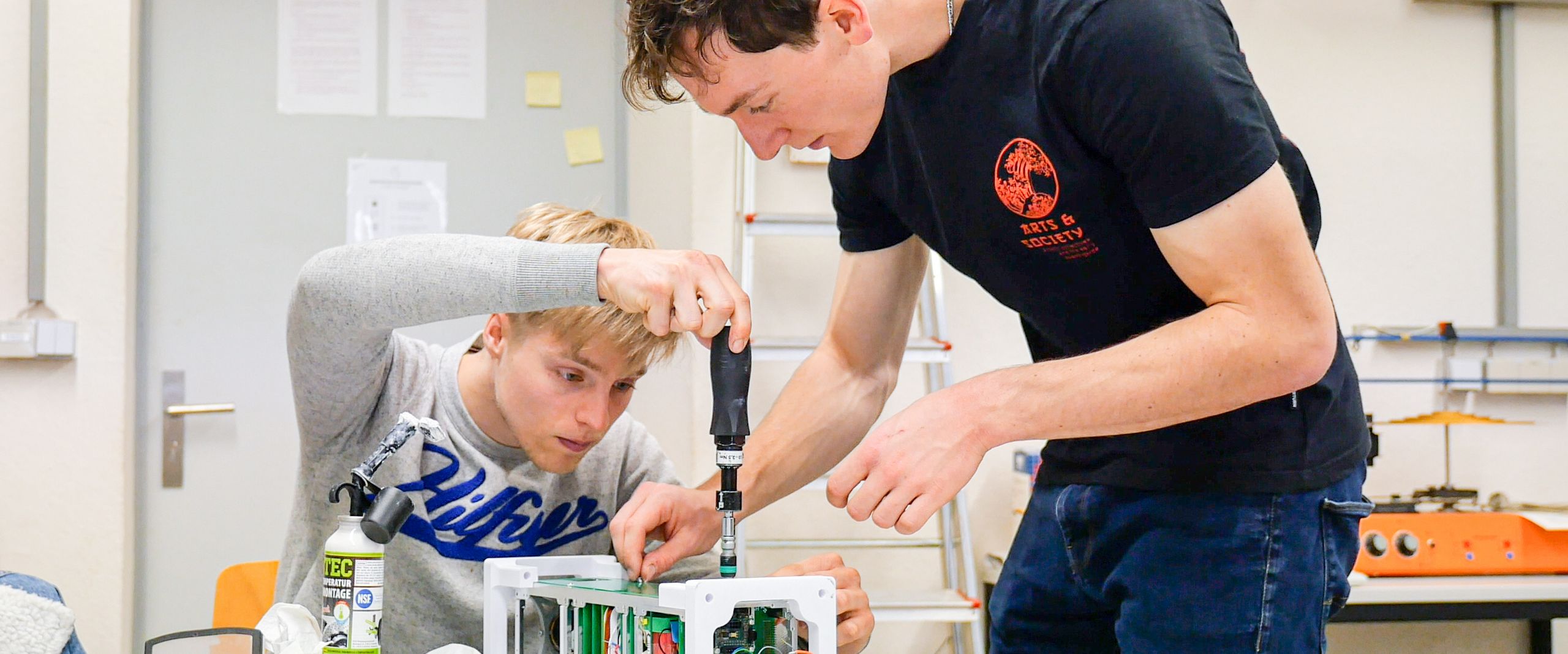 Zwei Studierende schrauben an einem technischen Gerät auf einem Tisch.