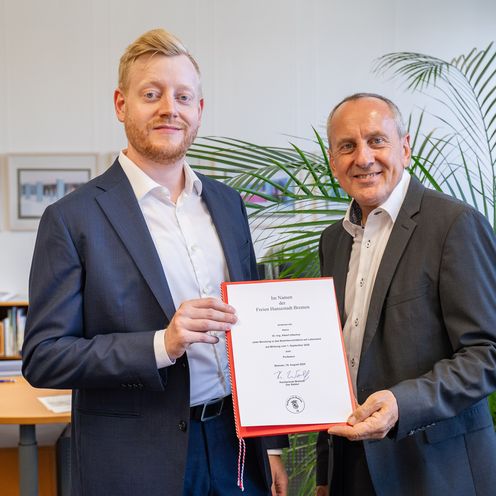 Zwei M&auml;nner halten gemeinsam eine Urkunde und schauen in die Kamera