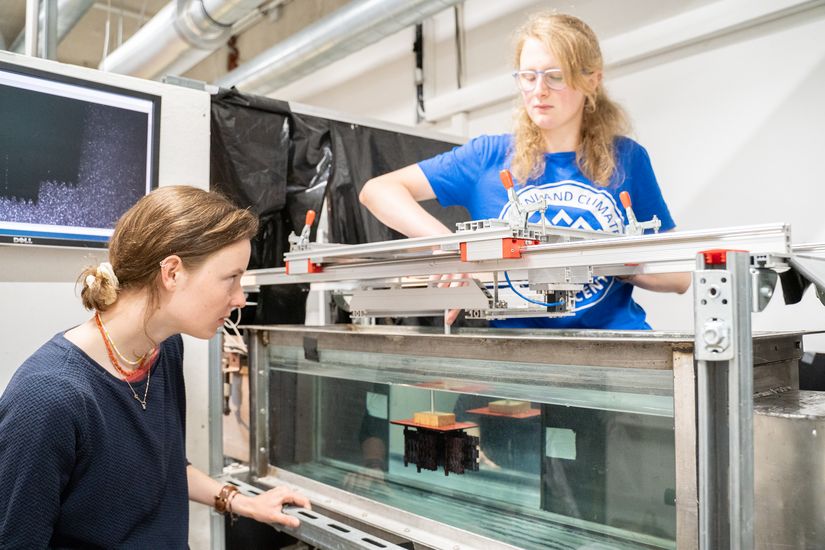 Zwei Studentinnen, die einander zugewandt vor und hinter einem Wasserkanal stehen. Eine Studentin h&auml;lt einen Probenk&ouml;rper ins Wasser. 