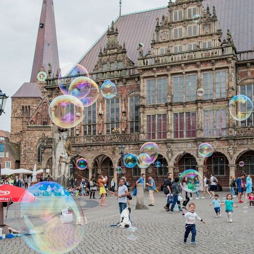 Auf dem Bild ist das Bremer Rathaus zu sehen. Im Vordergrund sind Seifenblasen, spielende Kinder und div. Menschen zu sehen. 