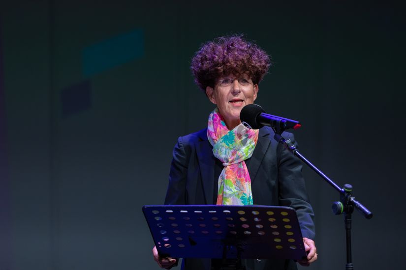 Frau mit buntem Schal steht an Rednerpult und Mikrofon und spricht