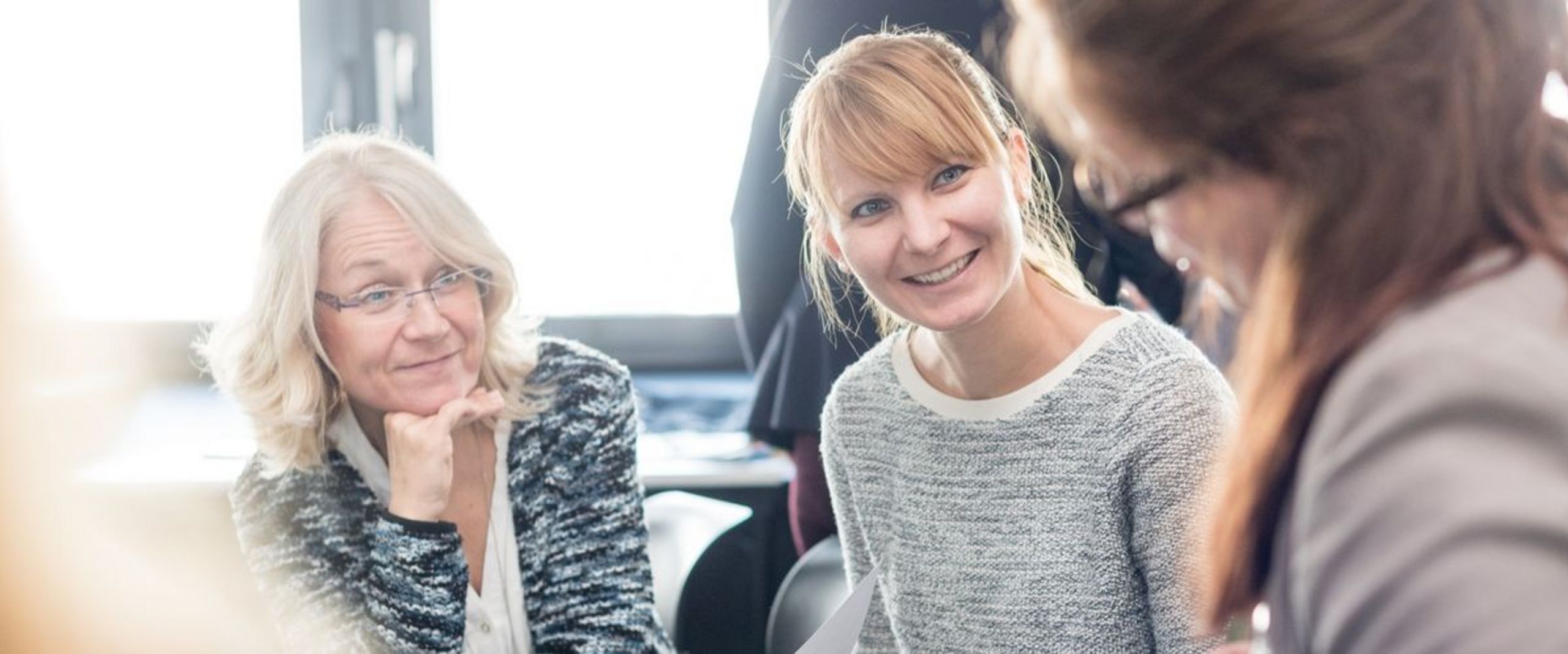 Zwei Frauen sitzen nebeneinander und sind im Fokus des Bildes. Eine stützt ihr Gesicht auf die Hand, eine hält ein Arbeitsblatt in der Hand. Beide lächeln und schauen eine dritte Frau an, die unscharf im Vordergrund des Bildes zu sehen ist. 