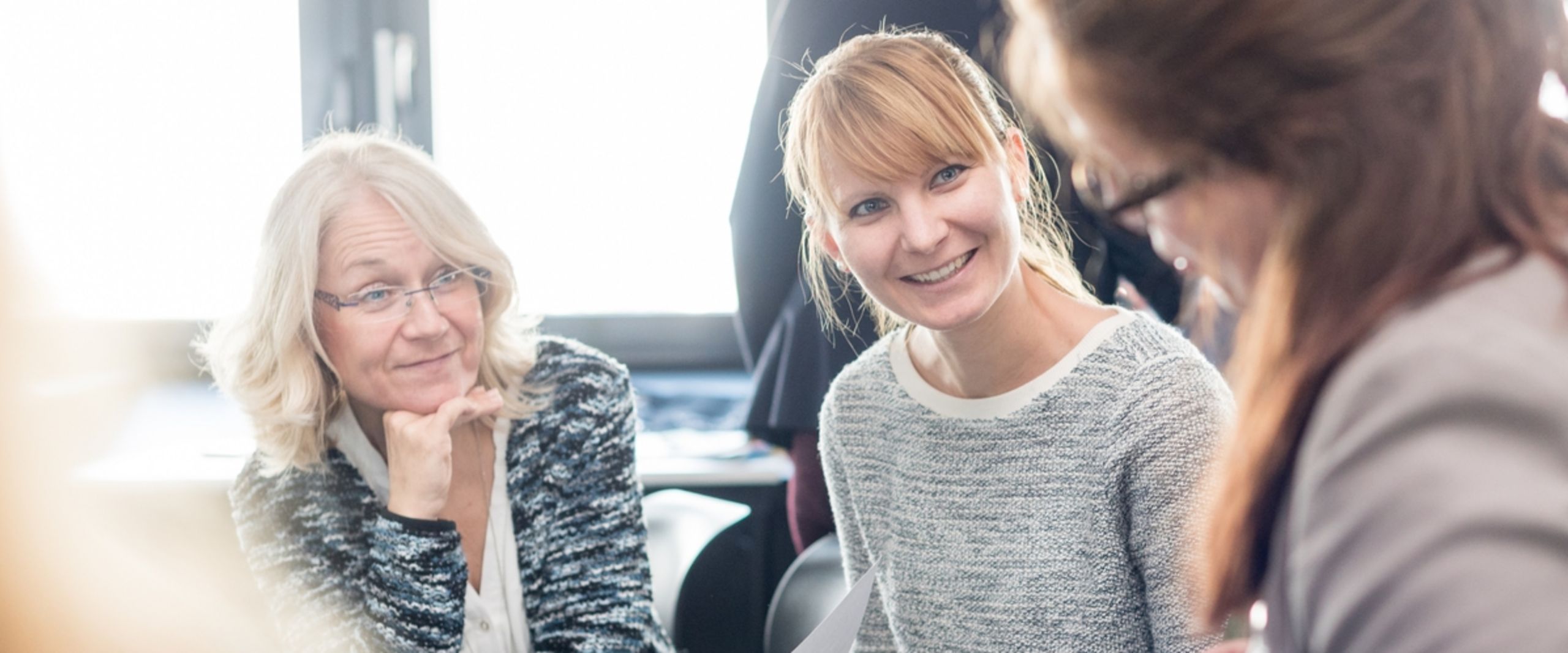 Kolleginnen im Gespräch Zwei Frauen sitzen nebeneinander und sind im Fokus des Bildes. Eine stützt ihr Gesicht auf die Hand, eine hält ein Arbeitsblatt in der Hand. Beide lächeln und schauen eine dritte Frau an, die unscharf im Vordergrund des Bildes zu sehen ist.