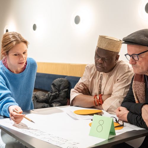 Das Bild zeigt eine Gruppe von Menschen mit diversem Hintergrund um einen Tisch miteinander sprechend. 