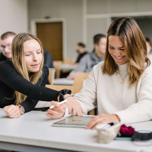 Zwei Studentinnen und ein Student sitzen an einem Tisch in einem Seminarraum. Im Hintergrund sieht man viele weitere Studierende. Die Studentin in der Mitte hat ein Tablet vor sich, einen Tablet-Stift in der Hand und lacht. Die Studentin neben ihr zeigt auf etwas auf dem Tablet. Der Student neben ihr schaut den beiden zu und lacht.