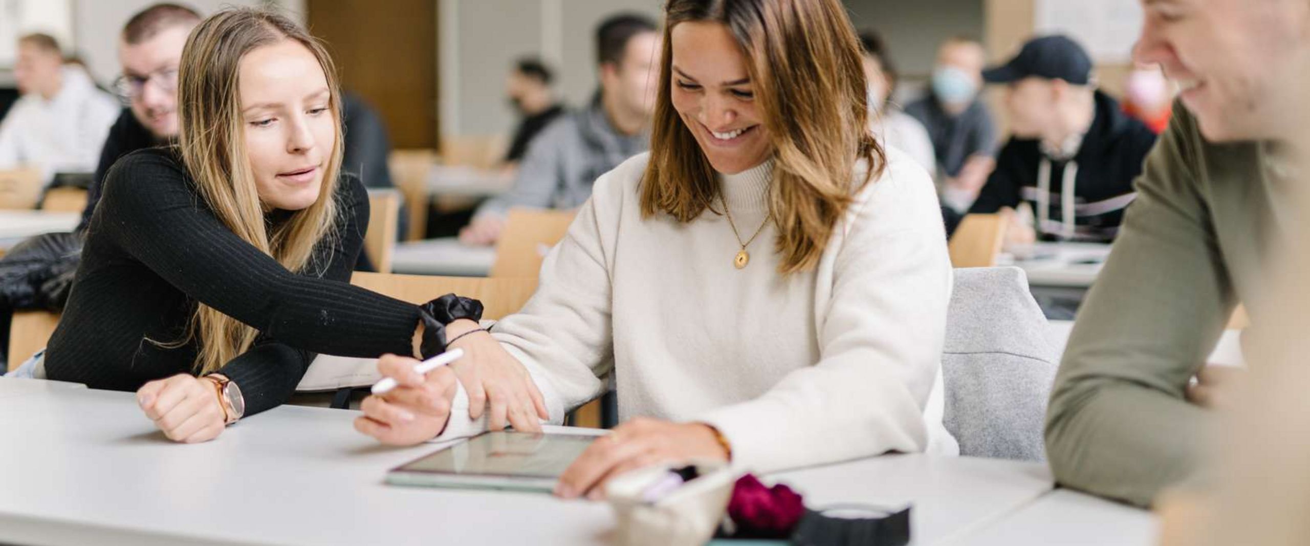 Zwei Studentinnen und ein Student sitzen an einem Tisch in einem Seminarraum. Im Hintergrund sieht man viele weitere Studierende. Die Studentin in der Mitte hat ein Tablet vor sich, einen Tablet-Stift in der Hand und lacht. Die Studentin neben ihr zeigt auf etwas auf dem Tablet. Der Student neben ihr schaut den beiden zu und lacht.