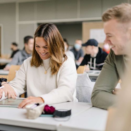 Zwei Studentinnen und ein Student sitzen an einem Tisch in einem Seminarraum. Im Hintergrund sieht man viele weitere Studierende. Die Studentin in der Mitte hat ein Tablet vor sich, einen Tablet-Stift in der Hand und lacht. Die Studentin neben ihr zeigt auf etwas auf dem Tablet. Der Student neben ihr schaut den beiden zu und lacht.