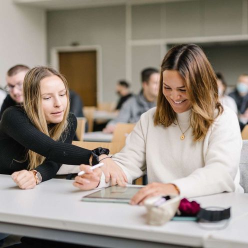 Zwei Studentinnen und ein Student sitzen an einem Tisch in einem Seminarraum. Im Hintergrund sieht man viele weitere Studierende. Die Studentin in der Mitte hat ein Tablet vor sich, einen Tablet-Stift in der Hand und lacht. Die Studentin neben ihr zeigt auf etwas auf dem Tablet. Der Student neben ihr schaut den beiden zu und lacht.