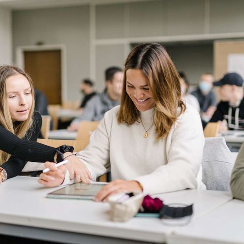 Studierende im Seminar Zwei Studentinnen und ein Student sitzen an einem Tisch in einem Seminarraum. Im Hintergrund sieht man viele weitere Studierende. Die Studentin in der Mitte hat ein Tablet vor sich, einen Tablet-Stift in der Hand und lacht. Die Studentin neben ihr zeigt auf etwas auf dem Tablet. Der Student neben ihr schaut den beiden zu und lacht.