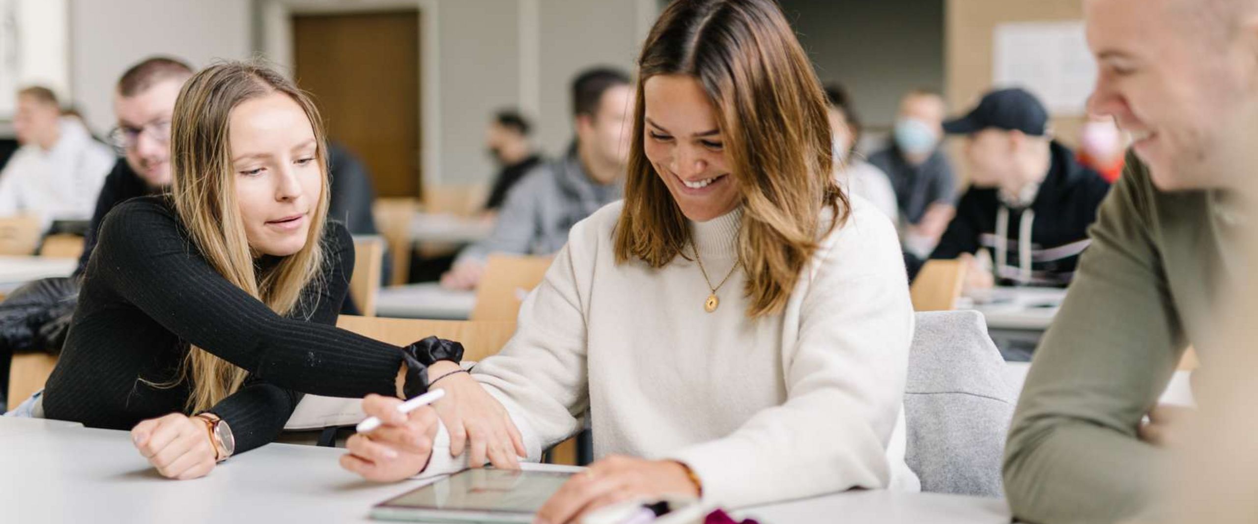 Zwei Studentinnen und ein Student sitzen an einem Tisch in einem Seminarraum. Im Hintergrund sieht man viele weitere Studierende. Die Studentin in der Mitte hat ein Tablet vor sich, einen Tablet-Stift in der Hand und lacht. Die Studentin neben ihr zeigt auf etwas auf dem Tablet. Der Student neben ihr schaut den beiden zu und lacht.