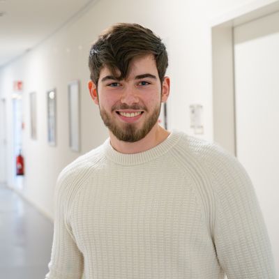 Auf dem Foto ist Moritz Lohff zusehen. Er hat dunkelbraune Haare und einen Bart und trägt einen weißen Strickpullover.