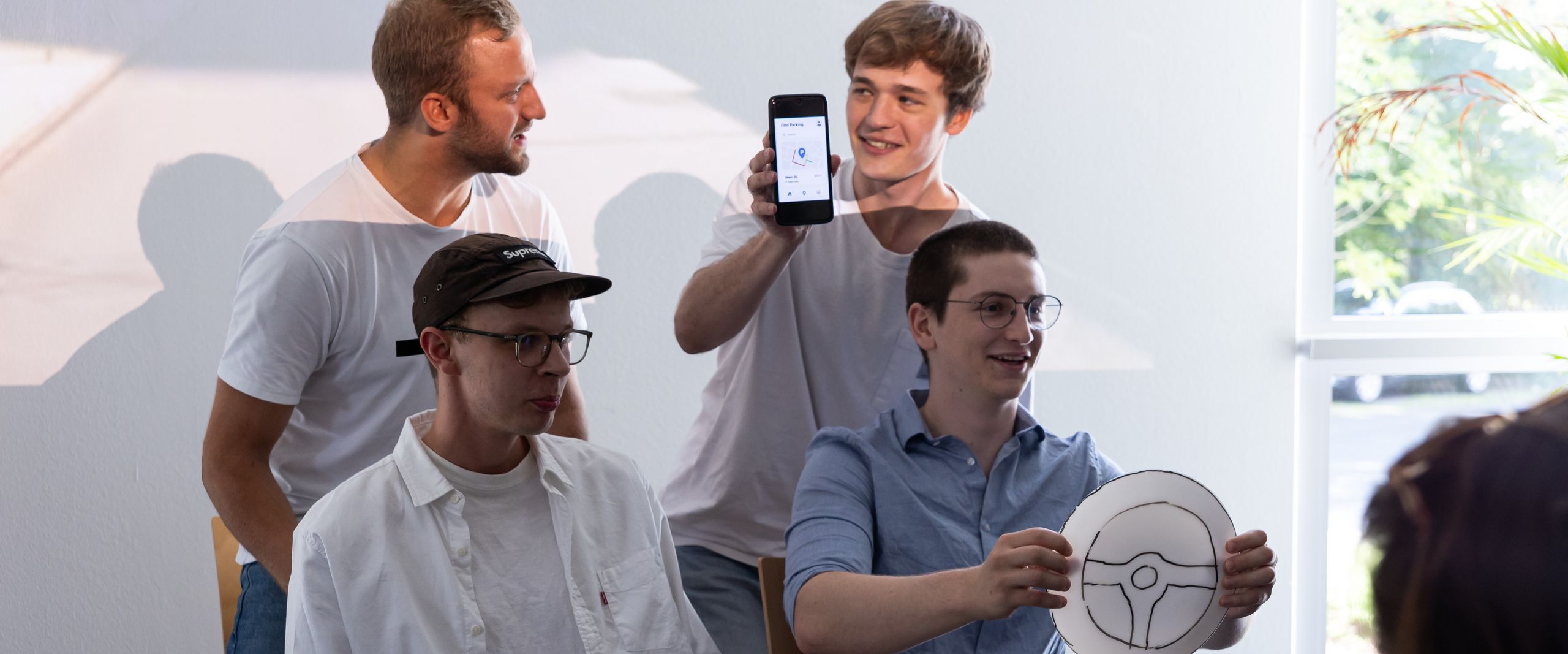 Startitup projektteam SPark 2025 Vier Männer und siitzen wie im Auto. der eine hält ein Lenkrad aus Papier.