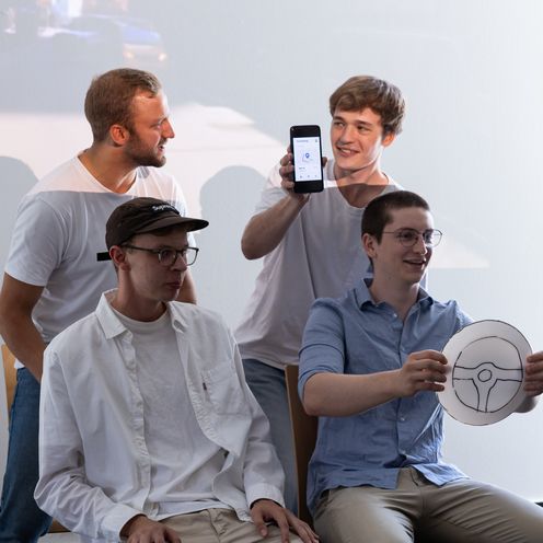 Vier M&auml;nner und siitzen wie im Auto. der eine h&auml;lt ein Lenkrad aus Papier.