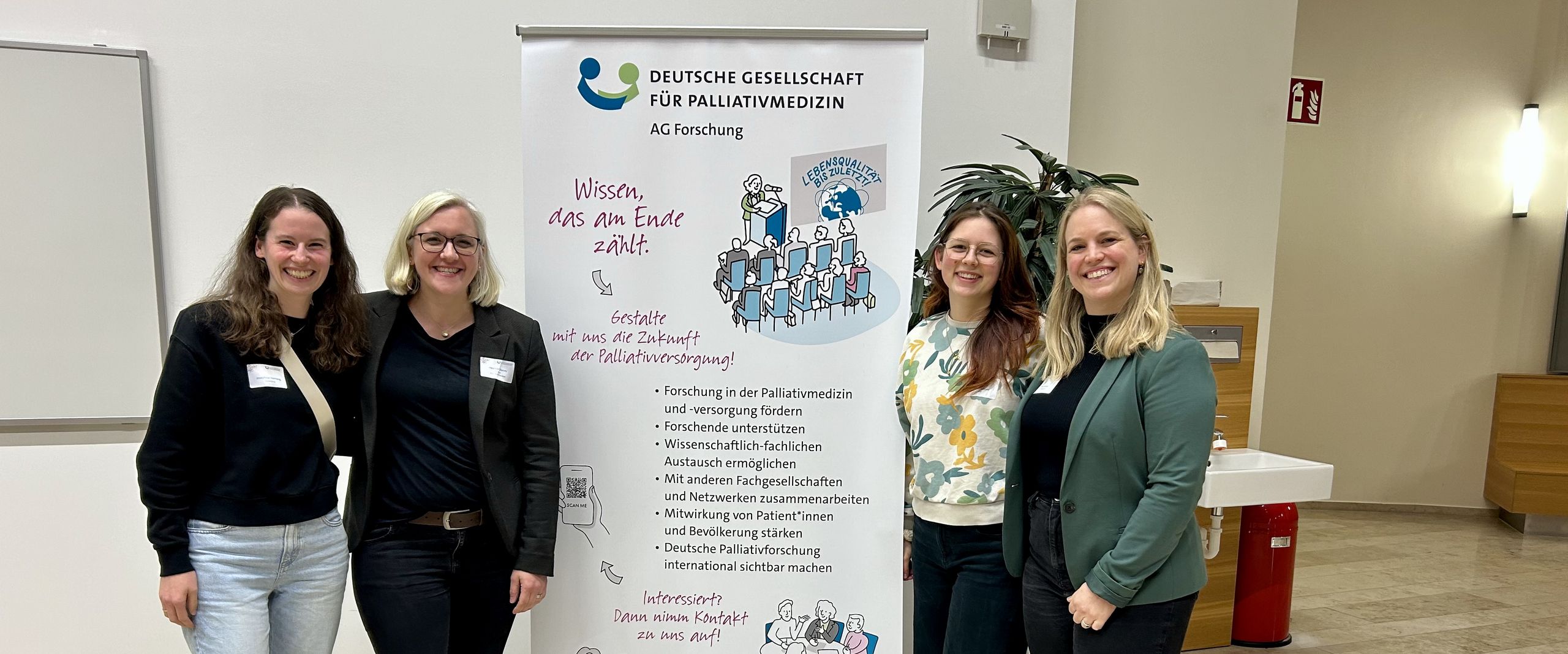 202503_Ausgezeichnet_Masterstudentinnen Palliativ Care Copyright Josephine Hornung Gruppenbild mit vier Frauen. In der Mitte ein Roll-up