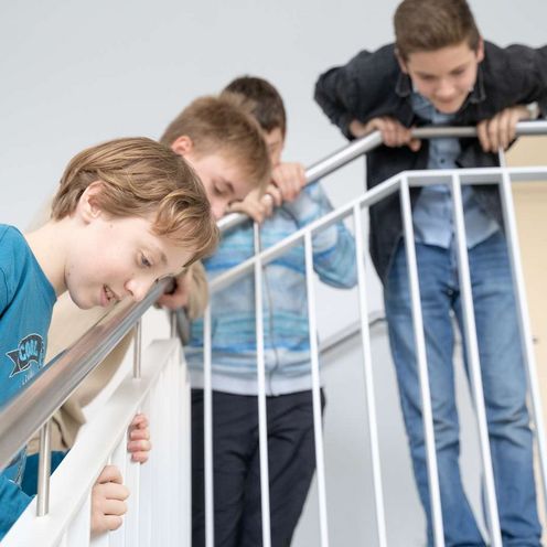 Auf dem Bild ist eine Gruppe von Sch&uuml;ler:innen zu sehen, die sich gespannt &uuml;ber das Treppengel&auml;nder nach unten beugen, um unten etwas zu beobachten.