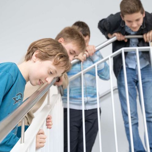 Auf dem Bild ist eine Gruppe von Sch&uuml;ler:innen zu sehen, die sich gespannt &uuml;ber das Treppengel&auml;nder nach unten beugen, um unten etwas zu beobachten.