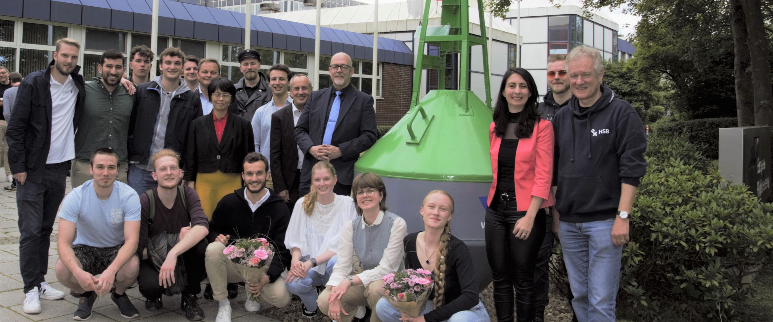 Gruppenbild mit Männern und Frauen an einer grünen Seefahrttonne. Im Hintergrund sieht man ein Gebäude mit Fahnen.