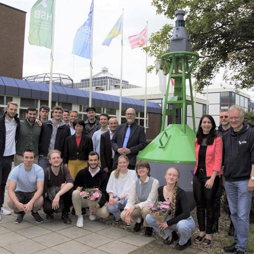 Gruppenbild mit M&auml;nnern und Frauen an einer gr&uuml;nen Seefahrttonne. Im Hintergrund sieht man ein Geb&auml;ude mit Fahnen.