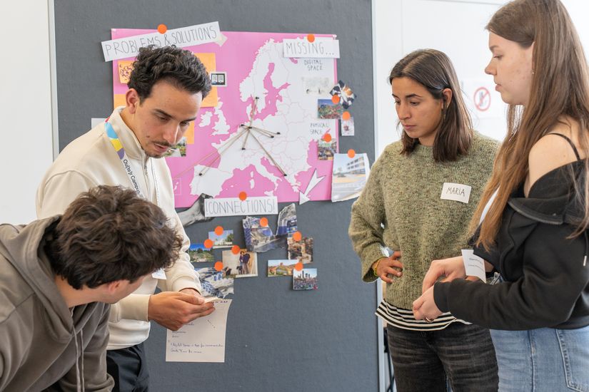 [Translate to English:] Eine Gruppe von Teilnehmenden arbeitet gemeinsam an einer Pinnwand mit einer Europakarte sowie Notizen und Bildern. Die Personen diskutieren Ideen und Verbindungen im Rahmen eines Workshops.