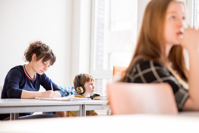 Studierende sitzt in einer Veranstaltung an einem tsich und schreibt. Neben der Person sitzt ein Kind mit Kopfhörern und blickt auf einen Laptop.