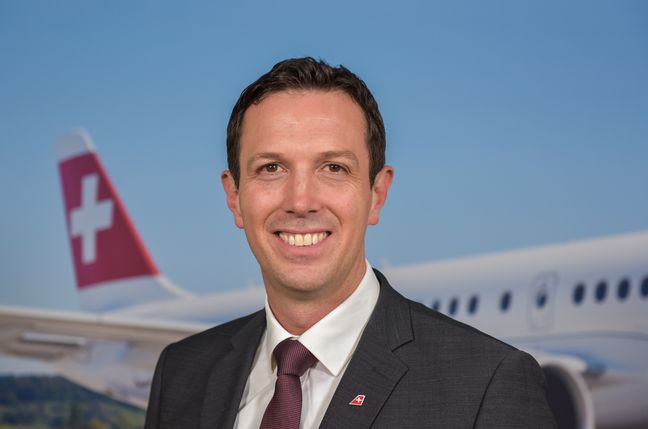 Jens Fehlender Auf dem Bild ist Jens Fehlender zusehen. er hat kurzes schwarzes Haar und trägt einen dunklen Anzug mit einer dunkel roten Krawatte. Im Hintergrund ist ein Flugzeug zu sehen.