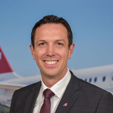 Auf dem Bild ist Jens Fehlender zusehen. er hat kurzes schwarzes Haar und trägt einen dunklen Anzug mit einer dunkel roten Krawatte. Im Hintergrund ist ein Flugzeug zu sehen. 