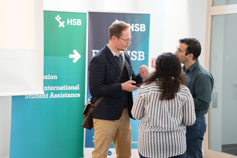 Drei Menschen stehen sich gegen&uuml;ber und unterhalten sich. Im Hintergrund h&auml;ngen HSB Plakate.