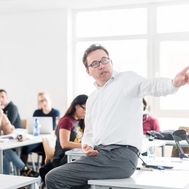 Studierende sitzen an Tischen in einem Seminarraum und hören einem Professor zu, der vor ihnen sitzt und etwas erklärt.