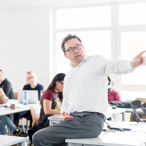 Studierende sitzen an Tischen in einem Seminarraum und h&ouml;ren einem Professor zu, der vor ihnen sitzt und etwas erkl&auml;rt.