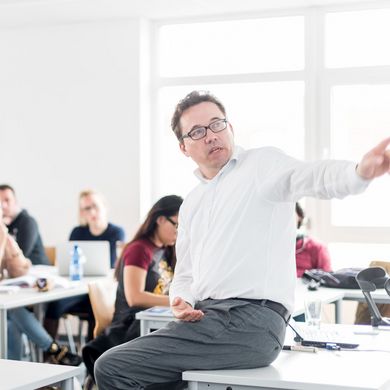Studierende sitzen an Tischen in einem Seminarraum und hören einem Professor zu, der vor ihnen sitzt und etwas erklärt.