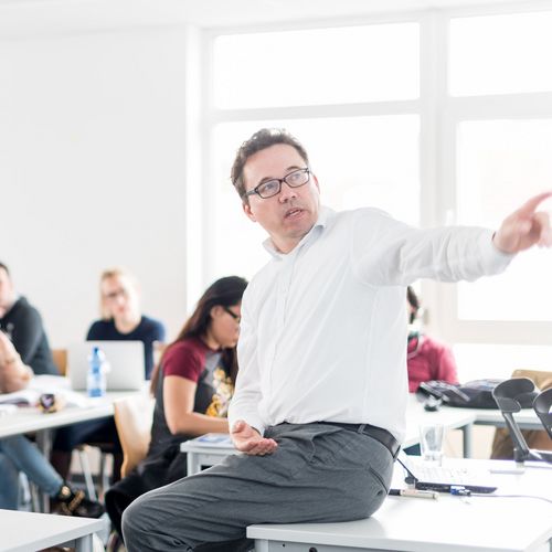 Studierende sitzen an Tischen in einem Seminarraum und h&ouml;ren einem Professor zu, der vor ihnen sitzt und etwas erkl&auml;rt.