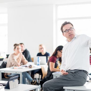 Studierende sitzen an Tischen in einem Seminarraum und hören einem Professor zu, der vor ihnen sitzt und etwas erklärt.