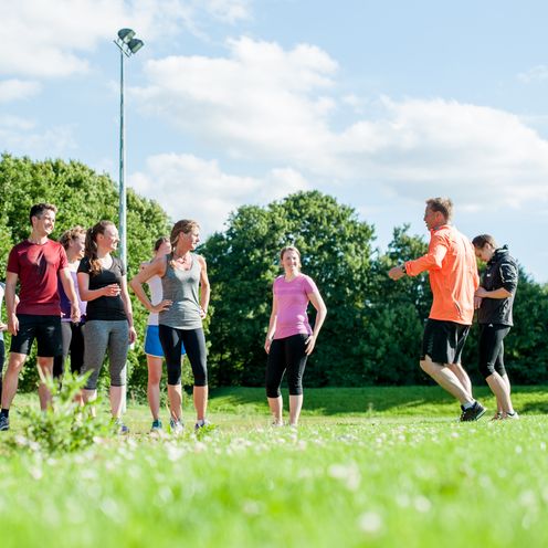 Eine Gruppe von Läufer:innen in bunter Sportkleidung steht auf einer Wiese und ist ihrem Trainer zugewandt, der auf der Stelle läuft.