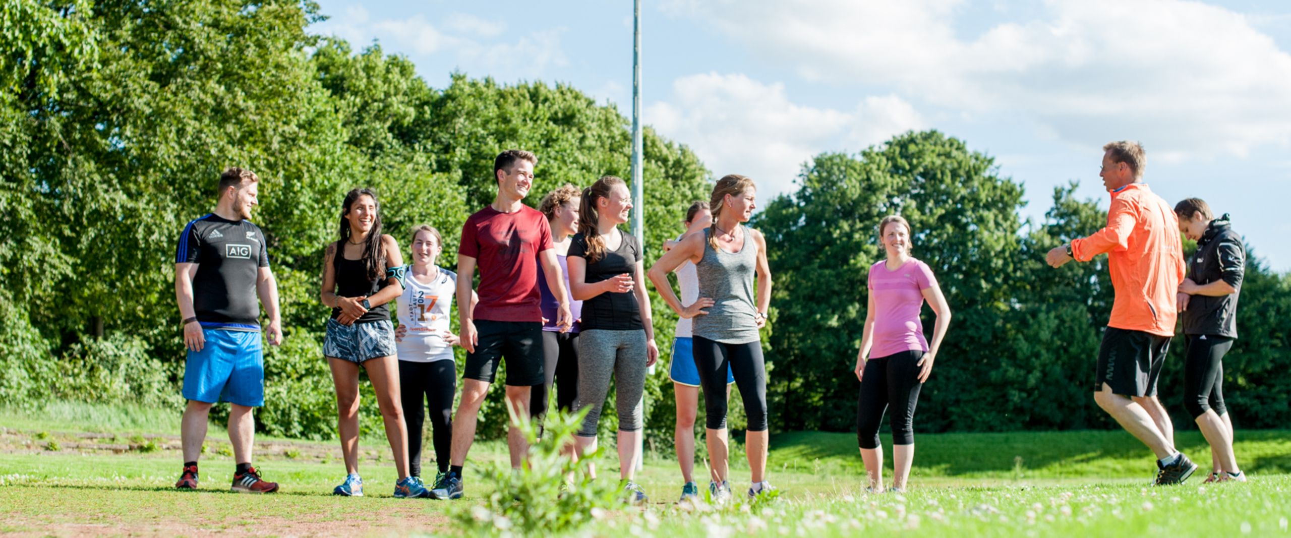 Eine Gruppe von Läufer:innen in bunter Sportkleidung steht auf einer Wiese und ist ihrem Trainer zugewandt, der auf der Stelle läuft.