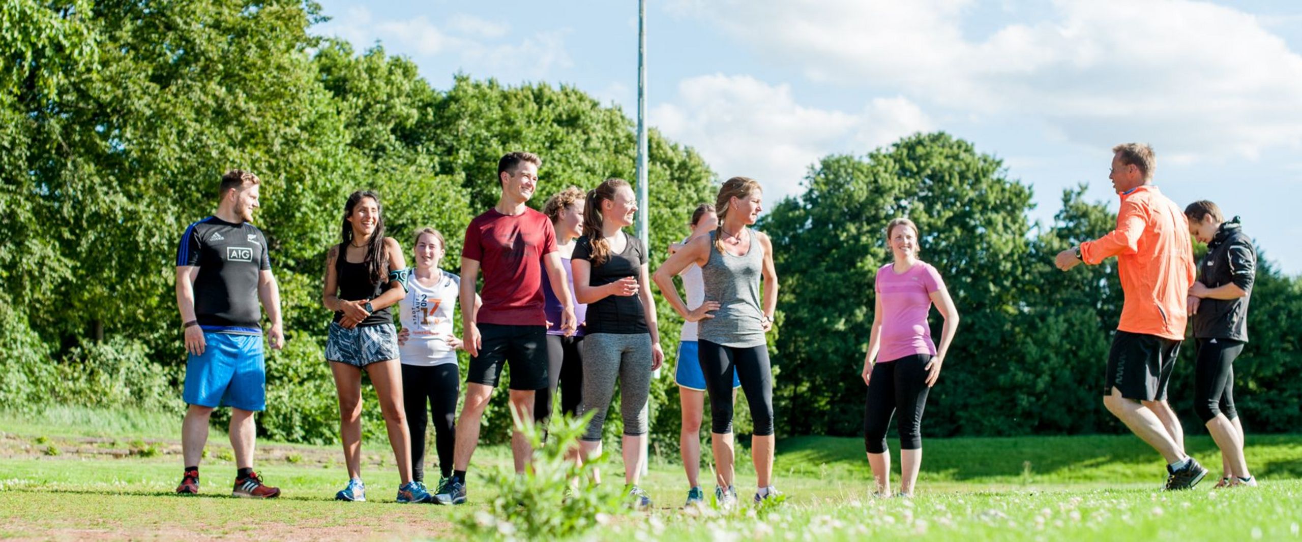 Eine Gruppe von Läufer:innen in bunter Sportkleidung steht auf einer Wiese und ist ihrem Trainer zugewandt, der auf der Stelle läuft.