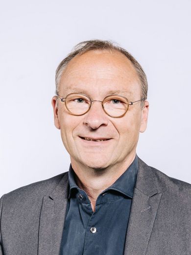 Auf dem Bild ist Rainer Hartmann zu sehen. Er hat kurzes graues Haar und eine braune runde Brille. Er trägt ein dunkel blaues Hemd unter einem grauen Jackett. 