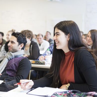 Studierende sitzen an Tischen in einer Vorlesung und lachen