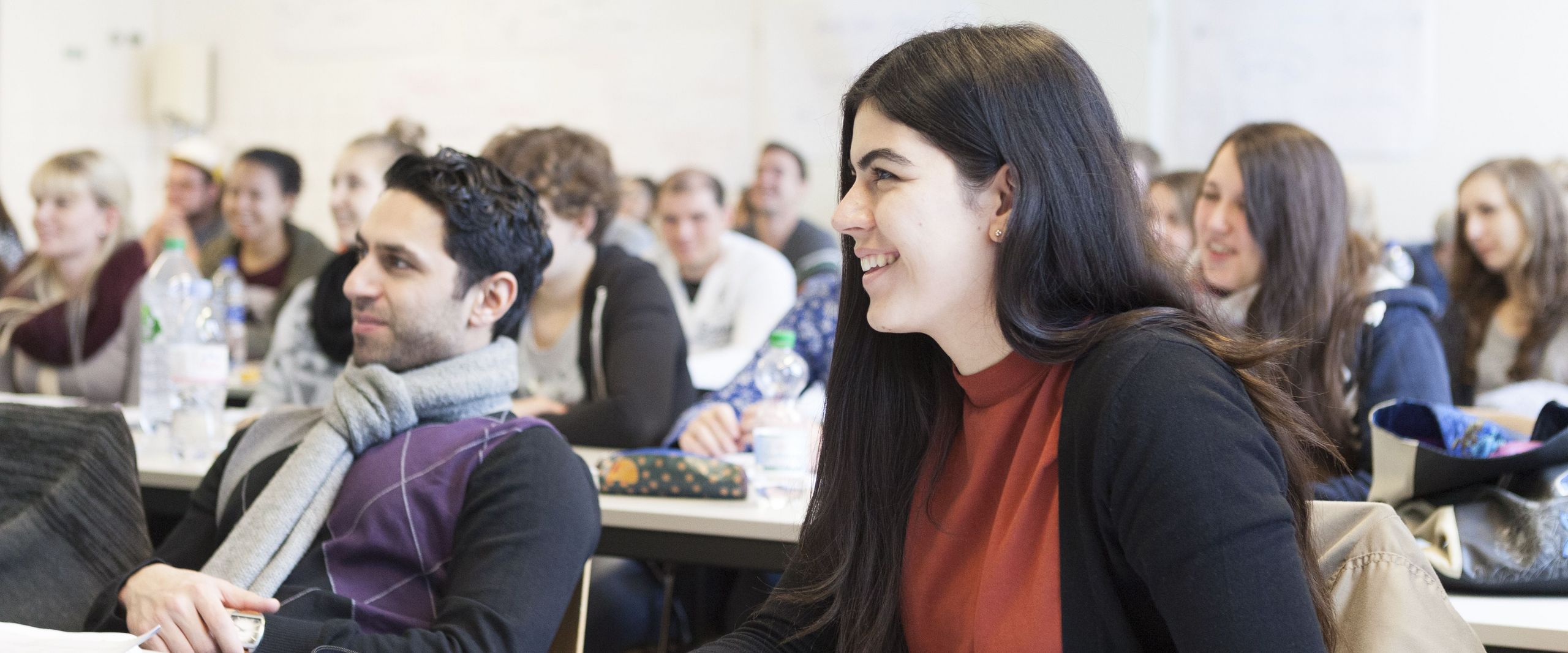 Studierende sitzen an Tischen in einer Vorlesung und lachen