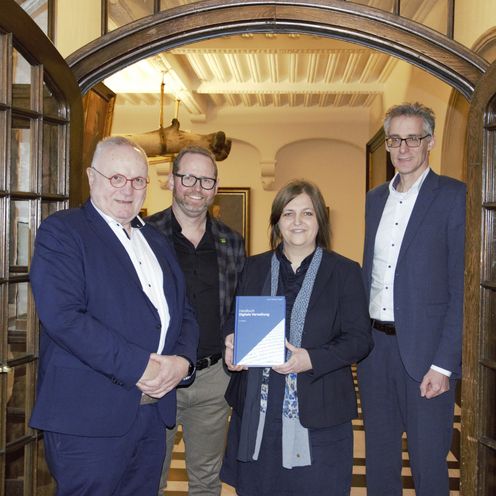 Gruppenbild mit drei M&auml;nnern und einer Frau. Diese pr&auml;sentiert in ihren H&auml;nden ein Buch. Alle stehen im Durchgang einer repr&auml;sentativen Holzt&uuml;r mit Glasfenstern. Beide T&uuml;rfl&uuml;gel sind weit ge&ouml;ffnet.