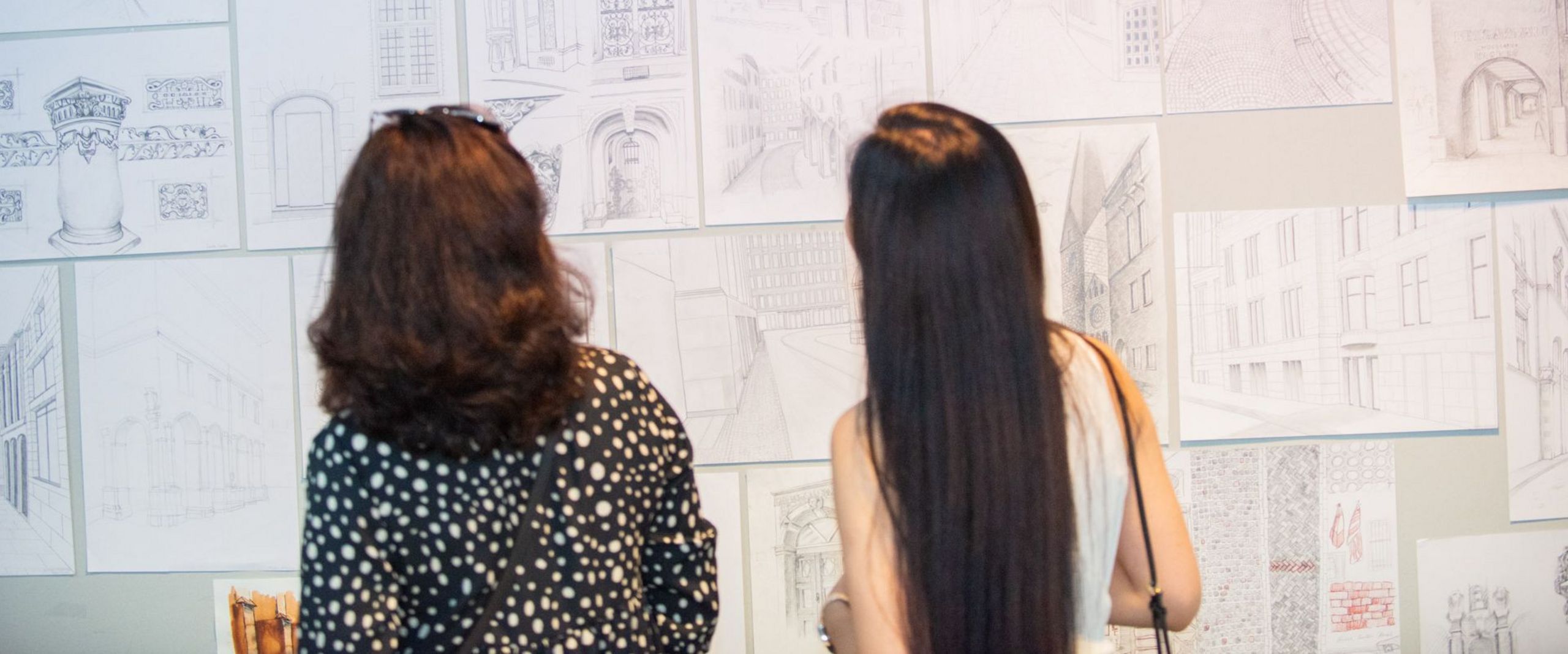 Zwei Frauen betrachten architektonische Skizzen an einer Wand. 