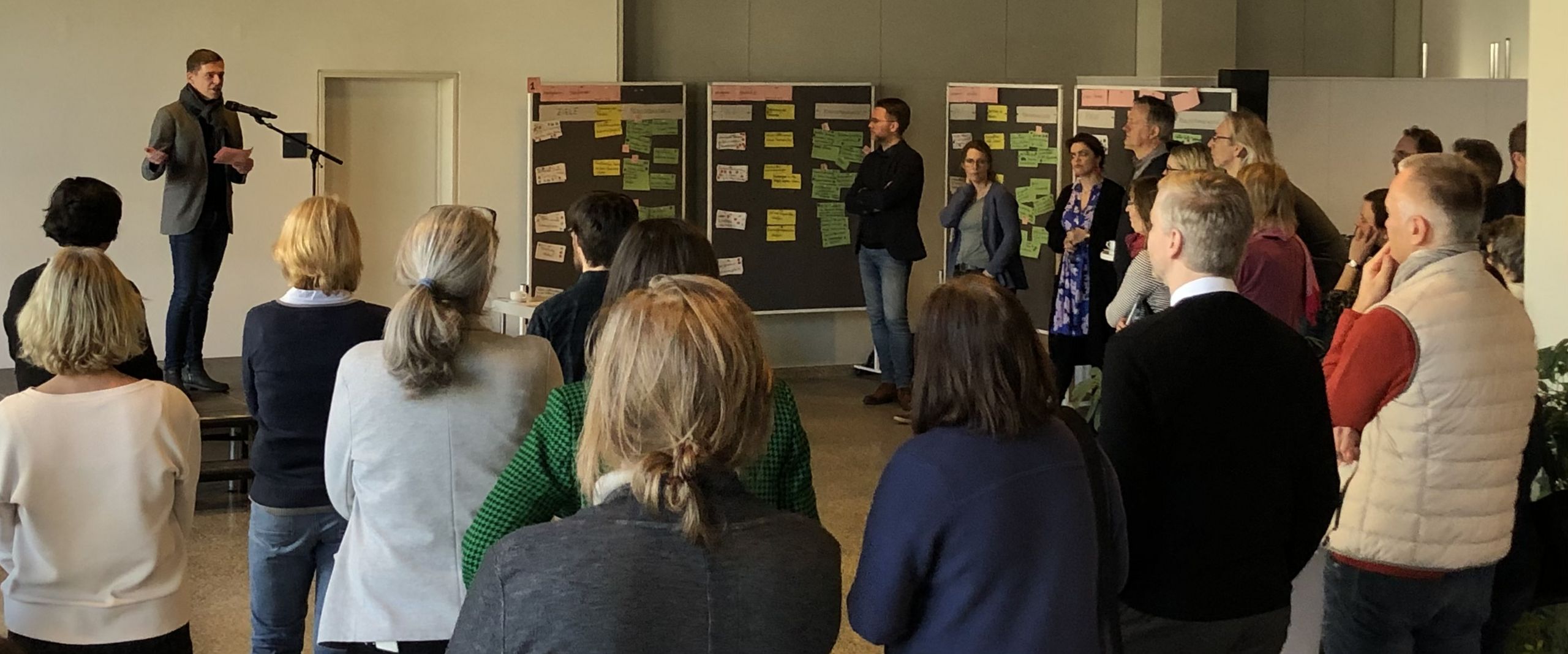 Menschen stehen in einem gro&szlig;en Raum zusammen und h&ouml;ren einem Mann auf der B&uuml;hne zu, der spricht. Rechts steht eine Pinnwand mit beschriebenen bunten Karten.