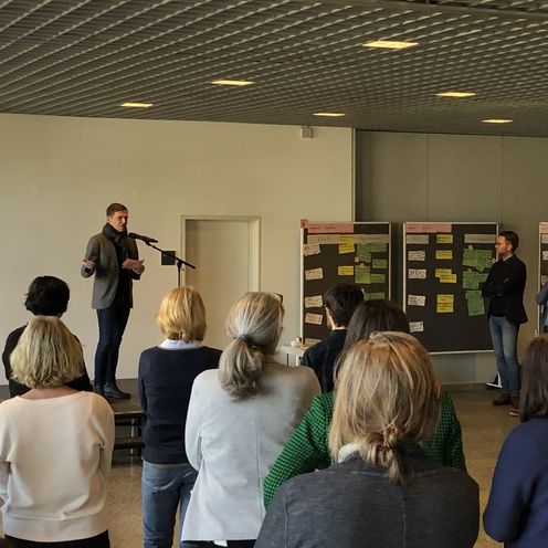 Menschen stehen in einem gro&szlig;en Raum zusammen und h&ouml;ren einem Mann auf der B&uuml;hne zu, der spricht. Rechts steht eine Pinnwand mit beschriebenen bunten Karten.