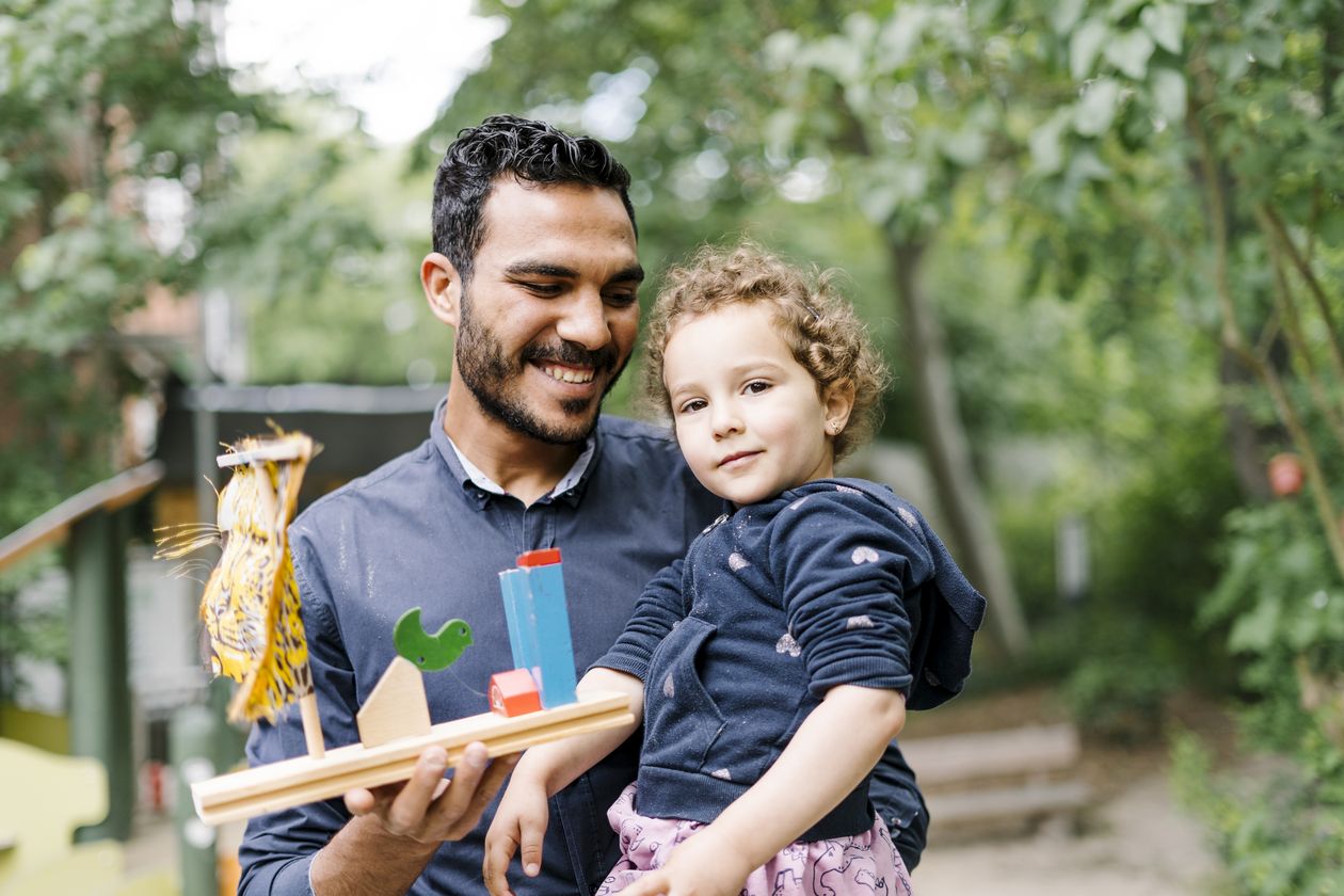 Ein Kleinkind auf dem Arm seines Elternteils. Das Elternteil hält ein gebasteltes Objekt in der anderen Hand.