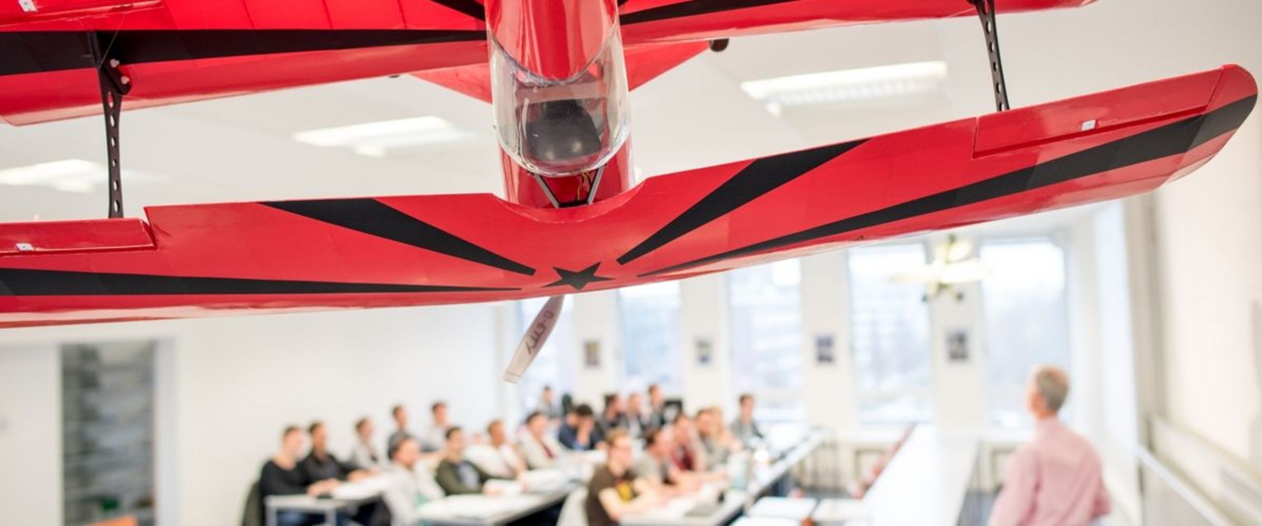 Auf dem Bild ist ein großes rotes Modellflugzeug zu sehen, das von der Decke eine Vorlesungssaales hängt. Im Hintergrund ist eine Grube Studierender und ein Lehrender im Seminarraum zu sehen.