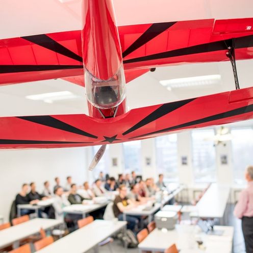 Auf dem Bild ist ein gro&szlig;es rotes Modellflugzeug zu sehen, das von der Decke eine Vorlesungssaales h&auml;ngt. Im Hintergrund ist eine Grube Studierender und ein Lehrender im Seminarraum zu sehen.