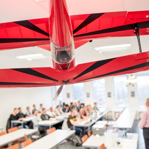 Auf dem Bild ist ein gro&szlig;es rotes Modellflugzeug zu sehen, das von der Decke eine Vorlesungssaales h&auml;ngt. Im Hintergrund ist eine Grube Studierender und ein Lehrender im Seminarraum zu sehen.